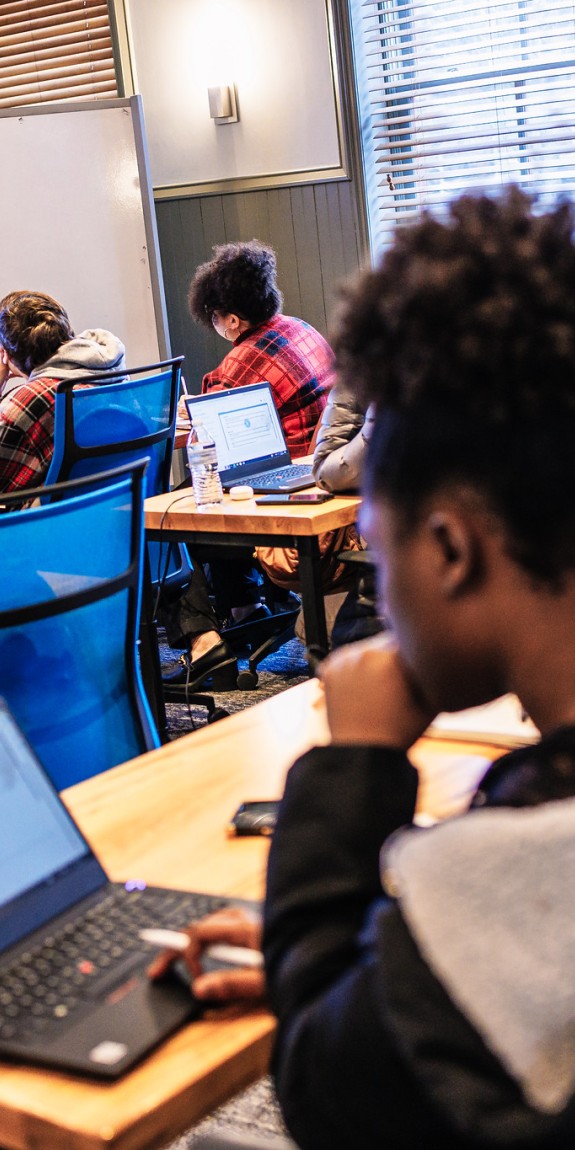 Students in a classroom working on laptops while a teacher instructs from a whiteboard. 